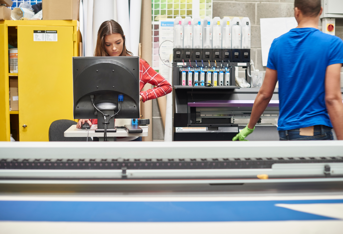 Two print shop workers operate digital label printing equipment, with one using a computer and the other working near a modern press.
