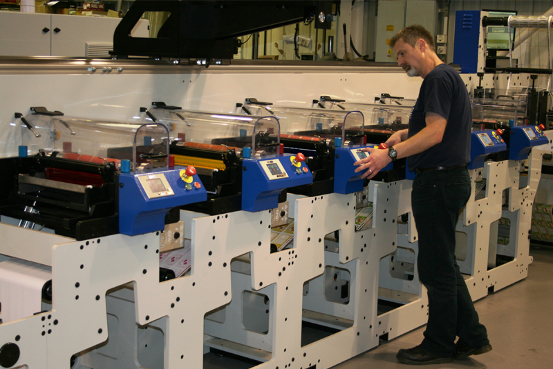 A man operates a flexo printing machine in an industrial setting, adjusting controls on a series of connected printing units with transparent covers.